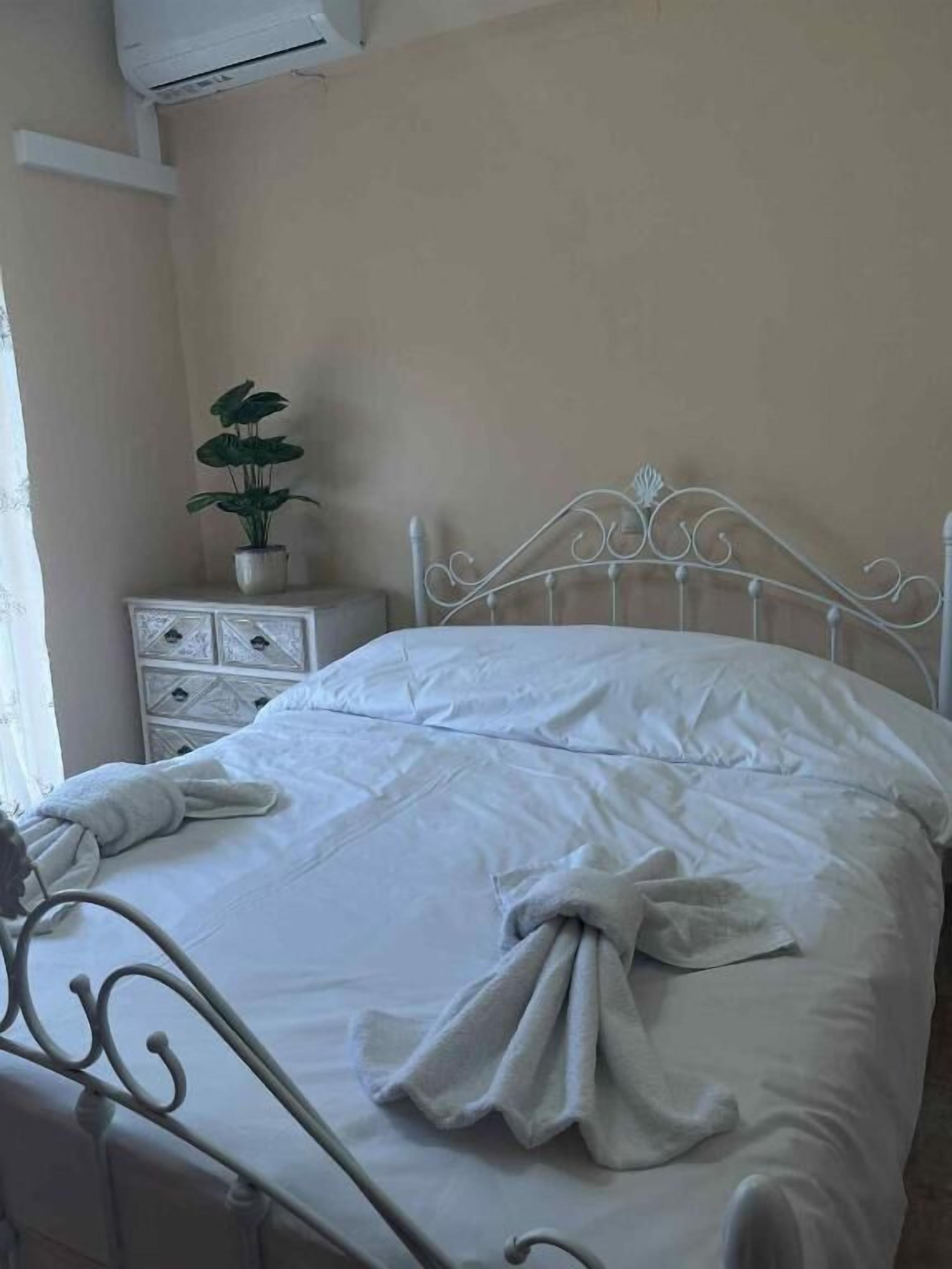 Uma cama bem arrumada com lençóis e toalha brancos, um vaso de planta em uma mesa de cabeceira e uma cabeceira de metal branca.