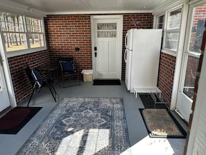 A covered porch with a brick wall, a white door, a refrigerator, and a blue patterned rug. - Privacy & space to unwind in this brick 3 bed home just minutes to the Beach! (Supply)