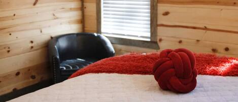 A wooden cabin room with a bed, a red blanket, and a black chair.