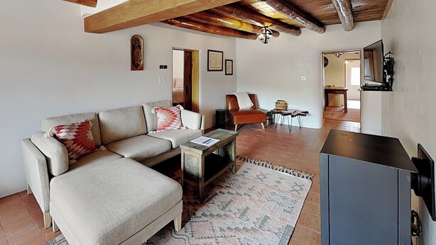 Una sala de estar con un sofá, una mesa de centro y una televisión. Hay una alfombra en el suelo y un techo con vigas a la vista.
