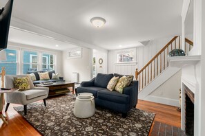 A living room with a blue sofa, a grey armchair, and a coffee table. There is a fireplace, a staircase, and a large window with blue shutters. - Holbrook House - Waterviews & Walk to Beach (Rockport)