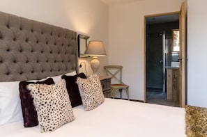 Ein Schlafzimmer mit einem gesteppten Kopfteil, einem Bett mit weißen Bettwäsche, leopardenmuster und dunkelbraunen Zierkissen, einer Nachttischlampe, einem Holzstuhl und einer Tür zu einem Küchenbereich.
