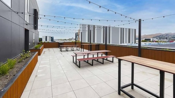 An outdoor patio with picnic tables, string lights, and a view of buildings and hills. - 2 Bedrooms Apprtmt Plus Underground Parking in CBD (Christchurch)