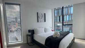 A modern hotel room with a large bed, a side table, a window with blinds, and a city view. - 2 Bedrooms Apprtmt Plus Underground Parking in CBD (Christchurch)