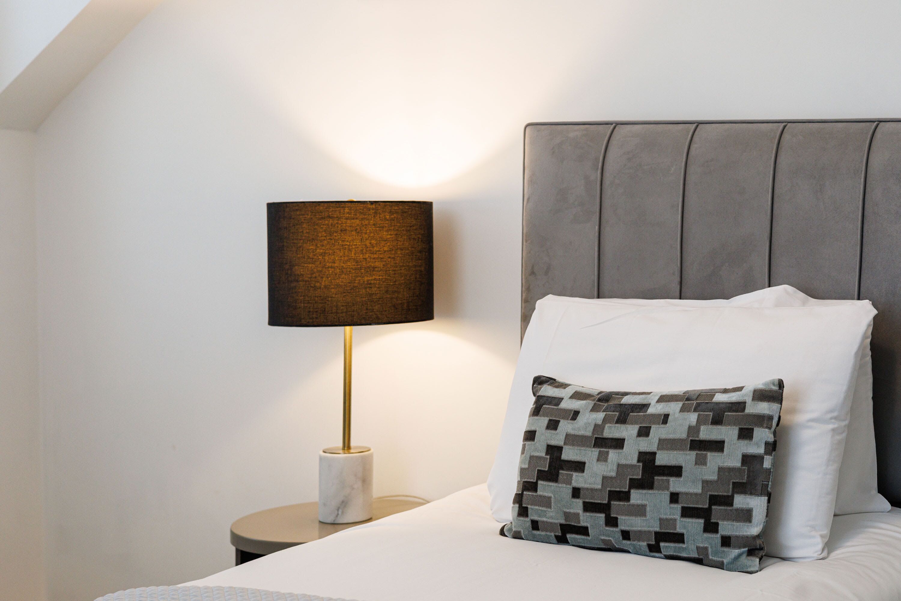 A neatly made bed with a grey upholstered headboard, a patterned pillow, and a lamp with a marble base on a bedside table.