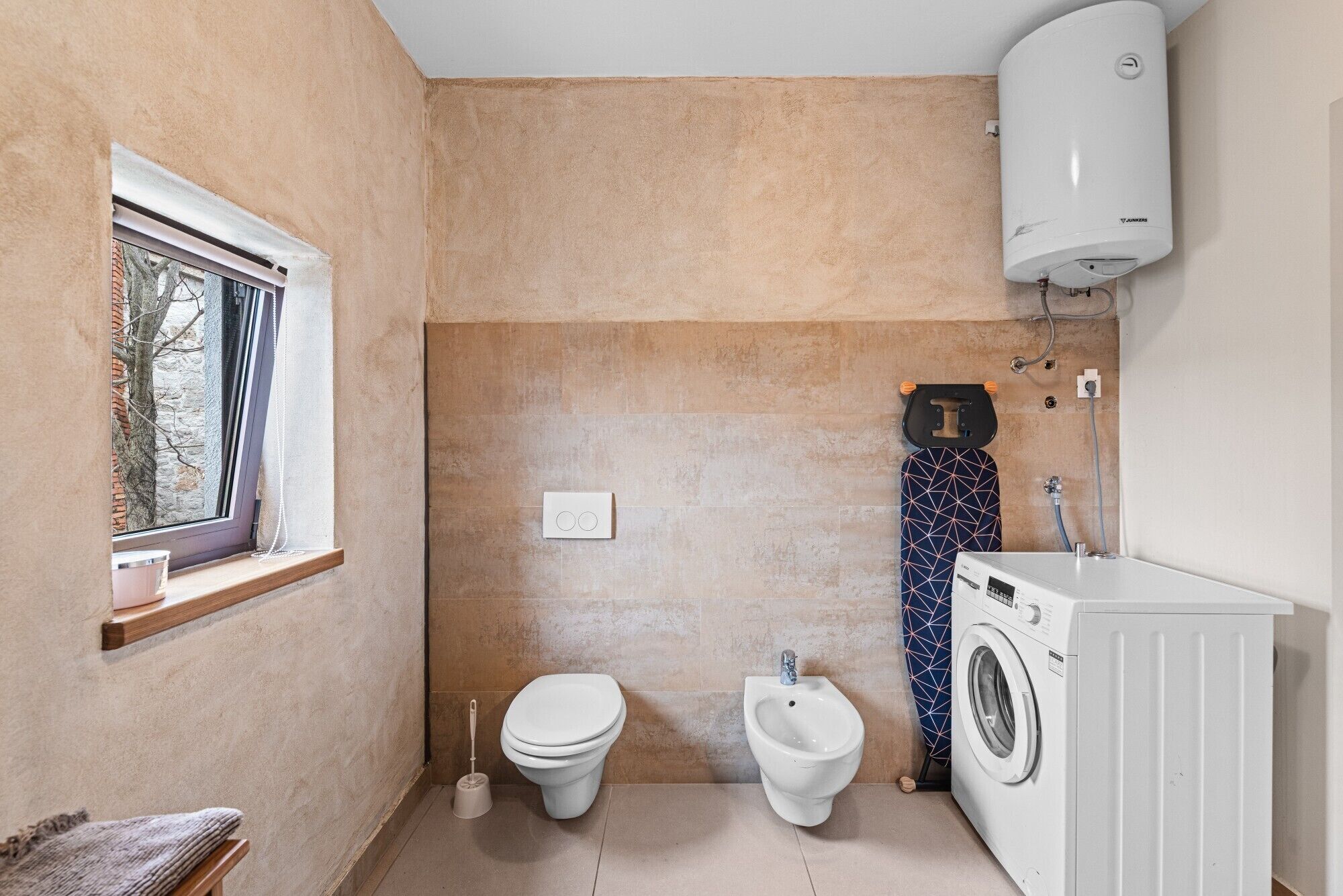 A compact bathroom with a toilet, bidet, washing machine, and a window.