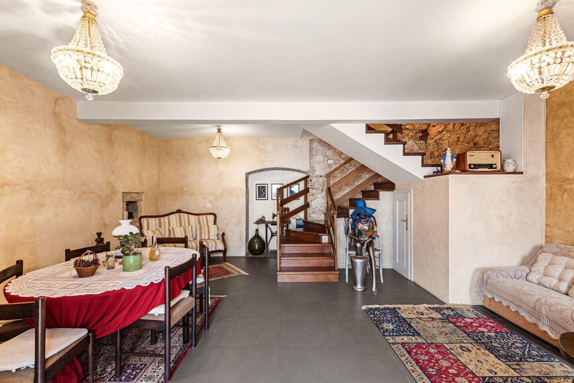 A dining area with a red tablecloth, wooden chairs, and a chandelier. There is a staircase leading to a loft area with a rustic stone wall.