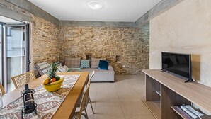Ein modernes Wohnzimmer mit einer Steinwand, einem Holzesstisch mit Obstschale, einer Couch und einem Fernseher auf einem Holzständer.