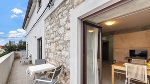 Ein Balkon mit Tisch und Stühlen, eine Steinmauer und eine Glastür zum Innenraum mit Fernseher und Esstisch.