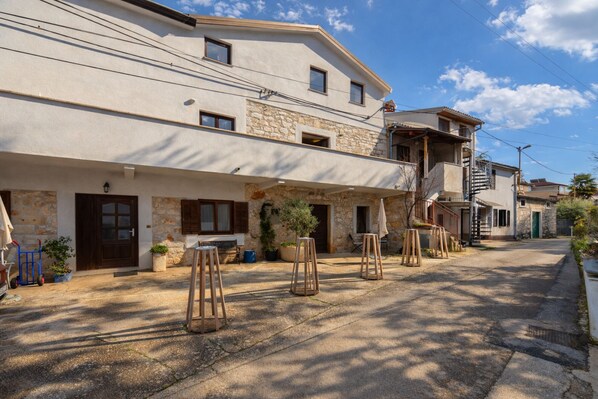 Un edificio de dos pisos con fachada de piedra, un balcón y una pequeña zona exterior con taburetes.