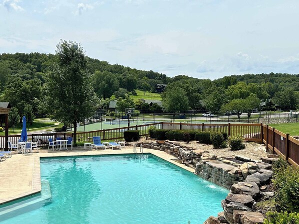 A swimming pool with a wooden deck and lounge chairs, surrounded by a wooden fence and a view of a tennis court and trees.