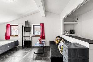 Ein gemütliches Zimmer mit Bett, Schreibtisch, Stuhl, einem kleinen Tisch mit Vase und einer Küchenzeile.