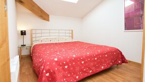 A bedroom with a wooden floor, a bed with a red bedspread, a nightstand with a lamp, and a white headboard. - Apartment Near Centre of Orelle (Orelle)