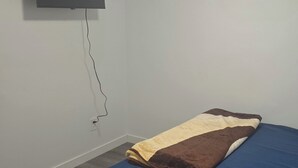 Une petite pièce, peu meublée, avec un lit simple, un téléviseur fixé au mur et un luminaire au plafond.