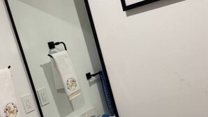 Une salle de bain avec un lavabo blanc, un miroir et des toilettes. Des serviettes sont accrochées au mur, un tableau encadré est placé au-dessus du miroir, et des articles de toilette sont posés sur le lavabo.