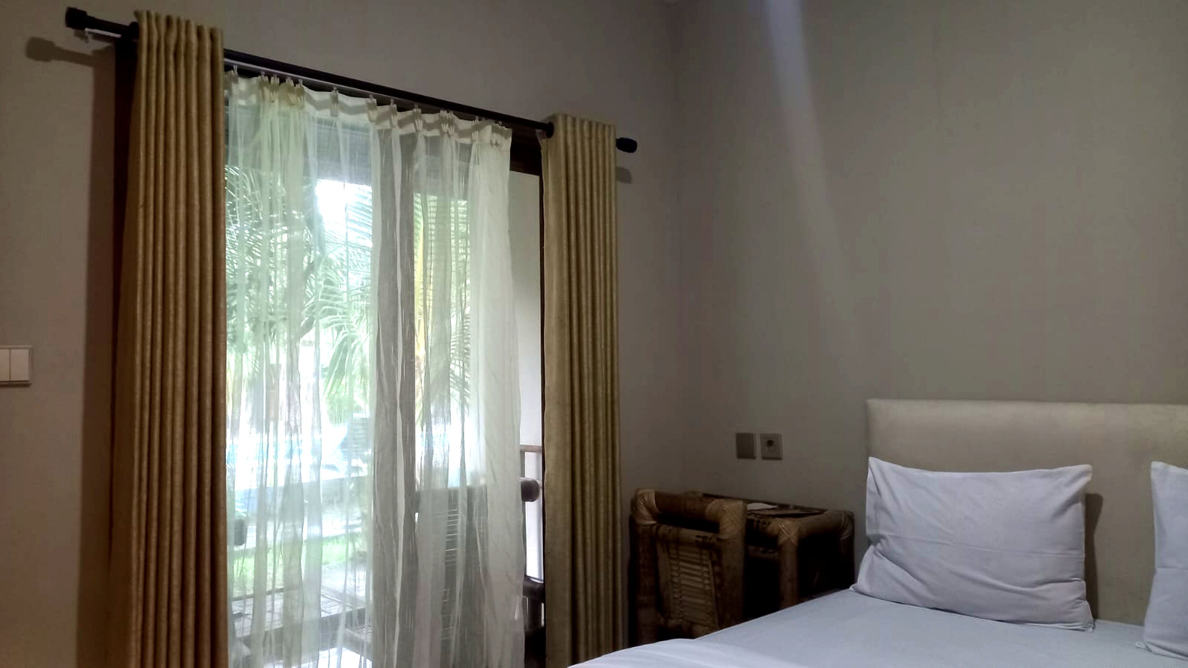 Hotelkamer met een bed, witte kussens, een hoofdbord en een raam met beige gordijnen.