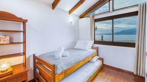 Un dormitorio con una cama de madera, una mesita de noche, una estantería con un objeto decorativo, y una ventana que ofrece vistas al mar y a las montañas.