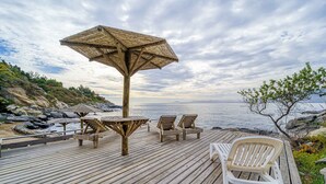 Una terraza de madera con una mesa, sillas, un parasol y vista al mar.
