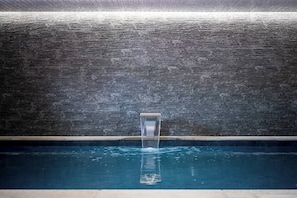 A swimming pool with a water feature in the center, surrounded by a dark brick wall. - Chalet Chêne - Panoramic Isere Valley Views (Sainte-Foy-Tarentaise)
