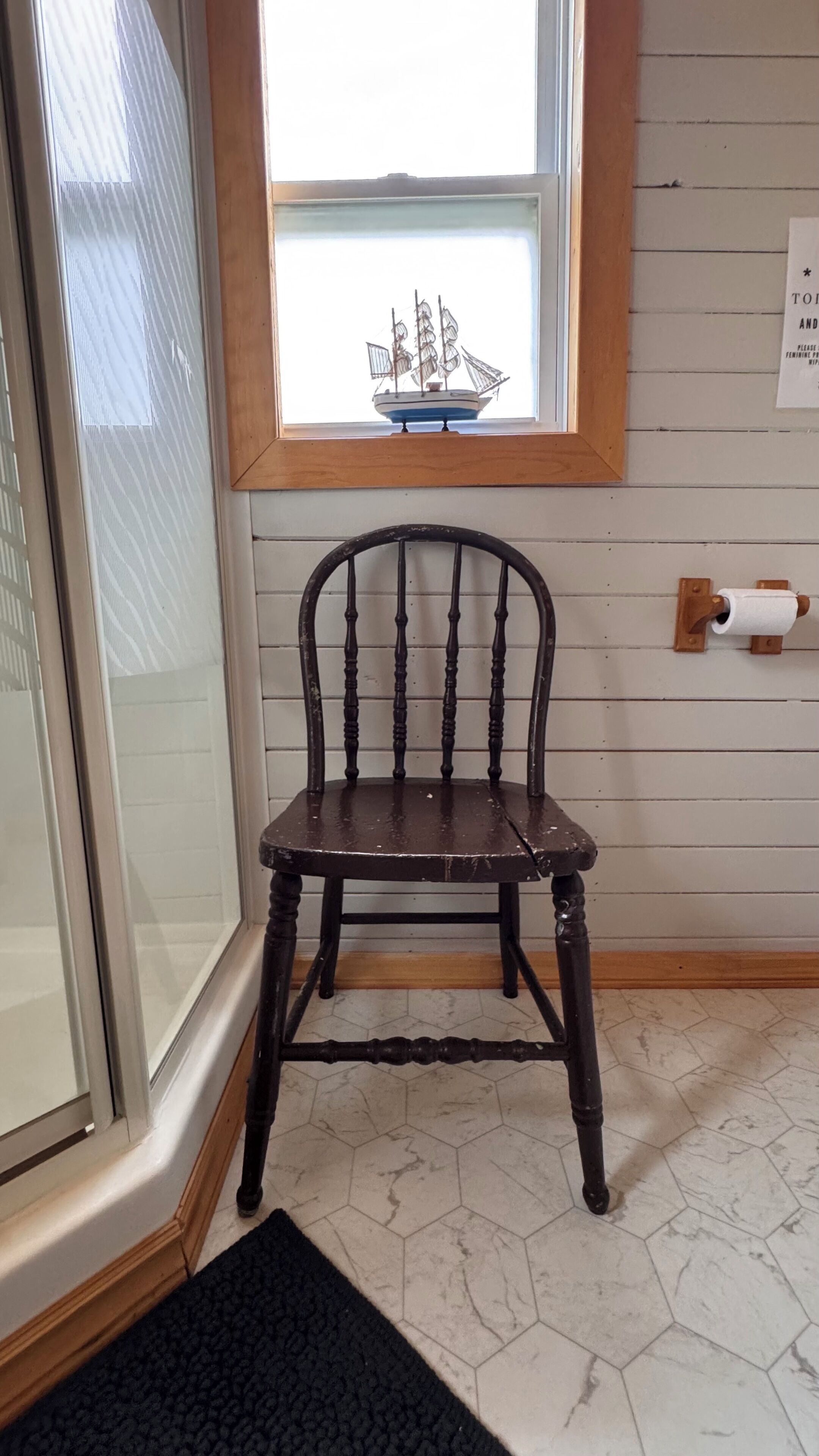 A wooden chair with a curved backrest and a dark finish, positioned in front of a window with a ship decoration.