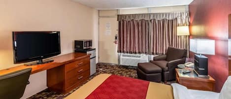 A hotel room with a bed, a desk with a television, a chair, and a bedside table with a lamp.