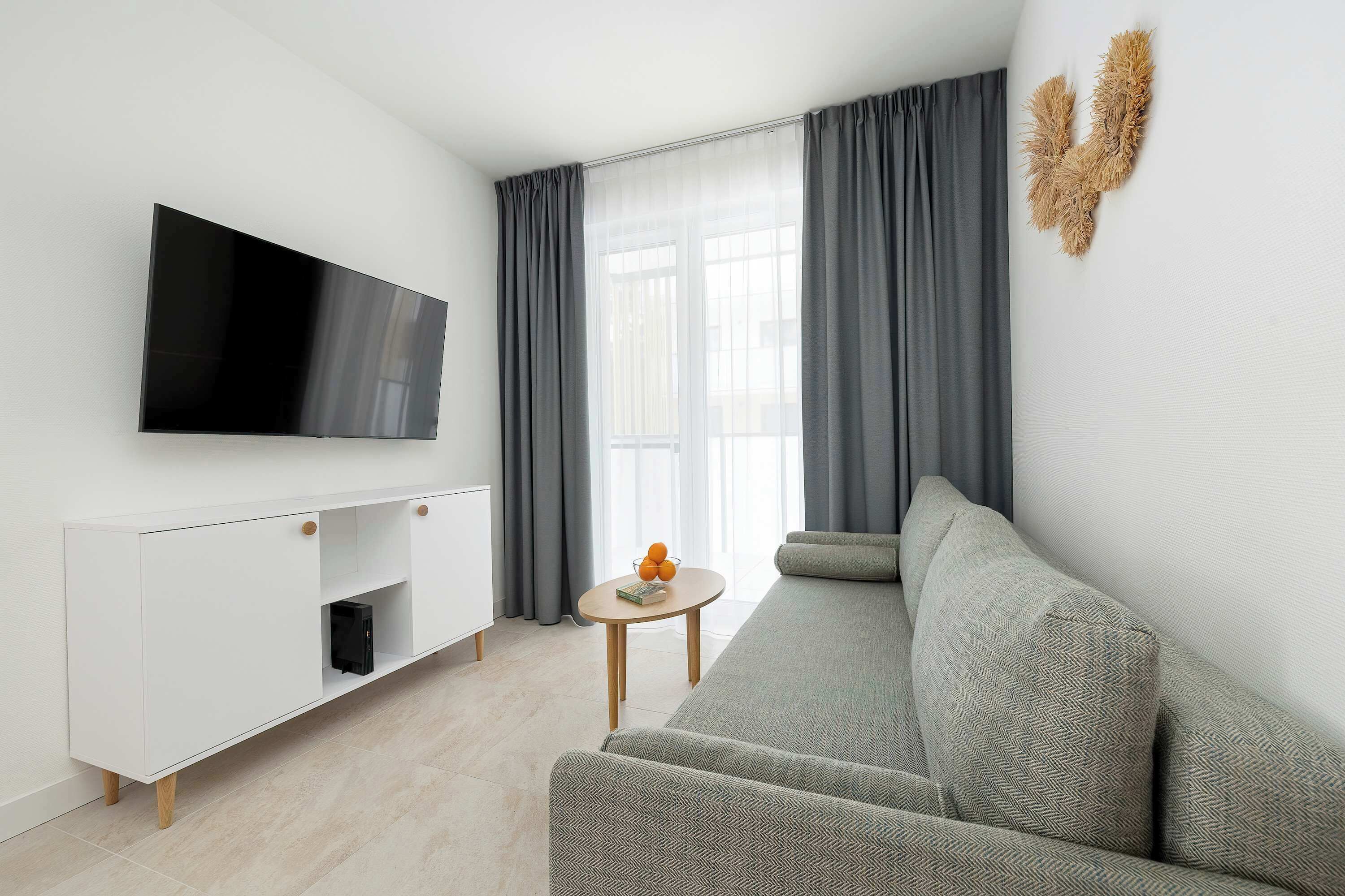 A bright living room features a comfortable grey sofa, a wooden coffee table, and a large flat-screen TV mounted above a white cabinet.