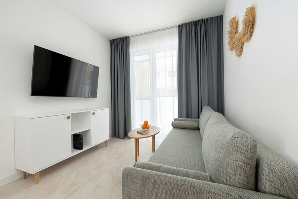 Ein modernes Wohnzimmer mit Flachbildfernseher, einer weißen Ablage, einem grauen Sofa und einem kleinen runden Tisch mit Obstschale.