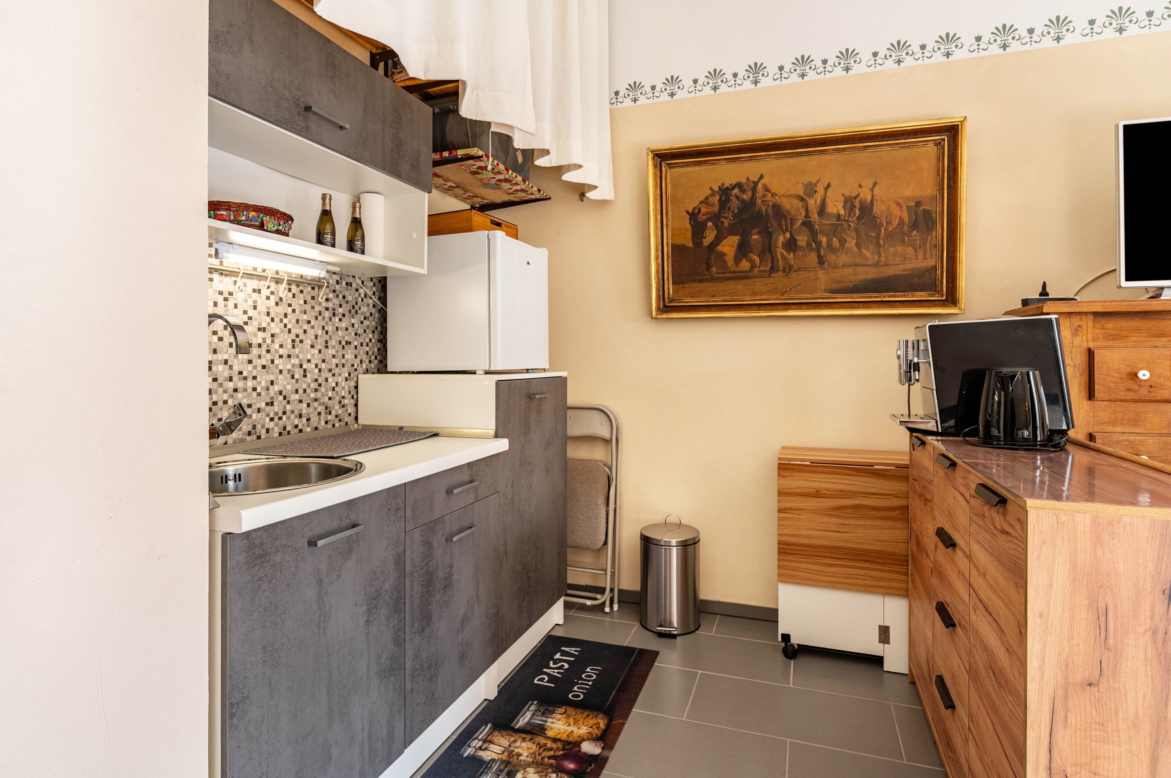 Een kleine keuken met een spoelbak, kastjes, een prullenbak, een koffiezetapparaat en een lijst aan de muur.