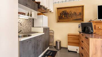 Een kleine keuken met een spoelbak, kastjes, een prullenbak, een koffiezetapparaat en een lijst aan de muur.