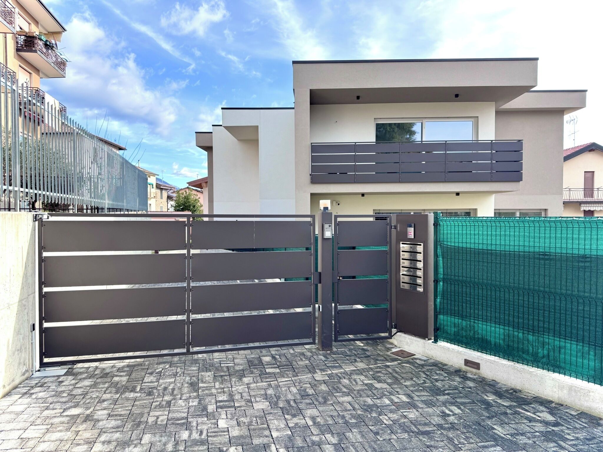 A gated entrance to a modern residential building with a paved driveway and a green fence.