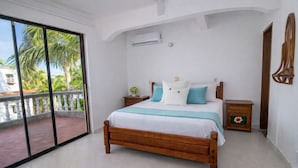 A bedroom with a wooden bed, a nightstand, a small cabinet, a balcony with a view of palm trees, and an air conditioning unit on the wall. - Ancla del galeon 7 (Tolú)