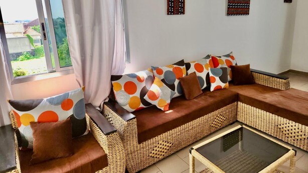 A living room with a wicker sofa, a glass coffee table, and a view of a garden through a window with curtains. - Private rooms in a Villa - Abidjan, Ivory Coast (Abidjan)