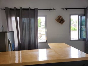 Una habitació amb taula de fusta, dues finestres amb cortines, prestatge fixat a la paret amb llibres i un frigorífic.