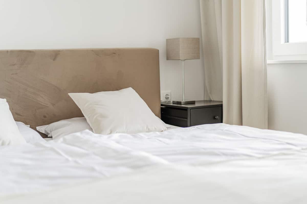 A neatly made bed with white linens, a bedside table with a lamp, and a window with curtains.