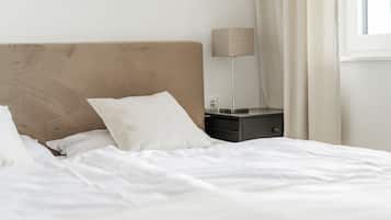 A neatly made bed with white linens, a bedside table with a lamp, and a window with curtains.