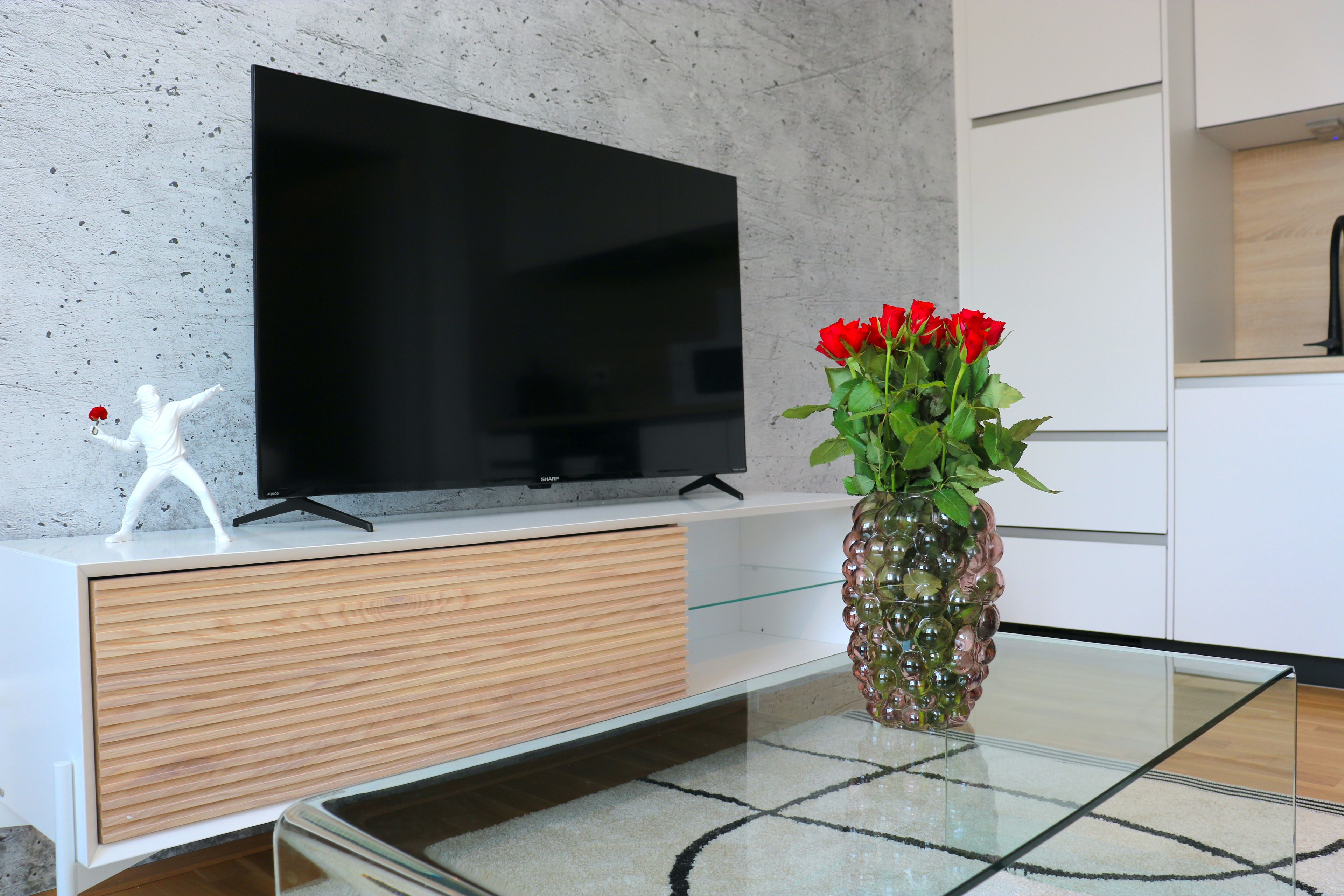 A modern living room with a flat-screen TV, a glass coffee table, and a vase with red flowers.