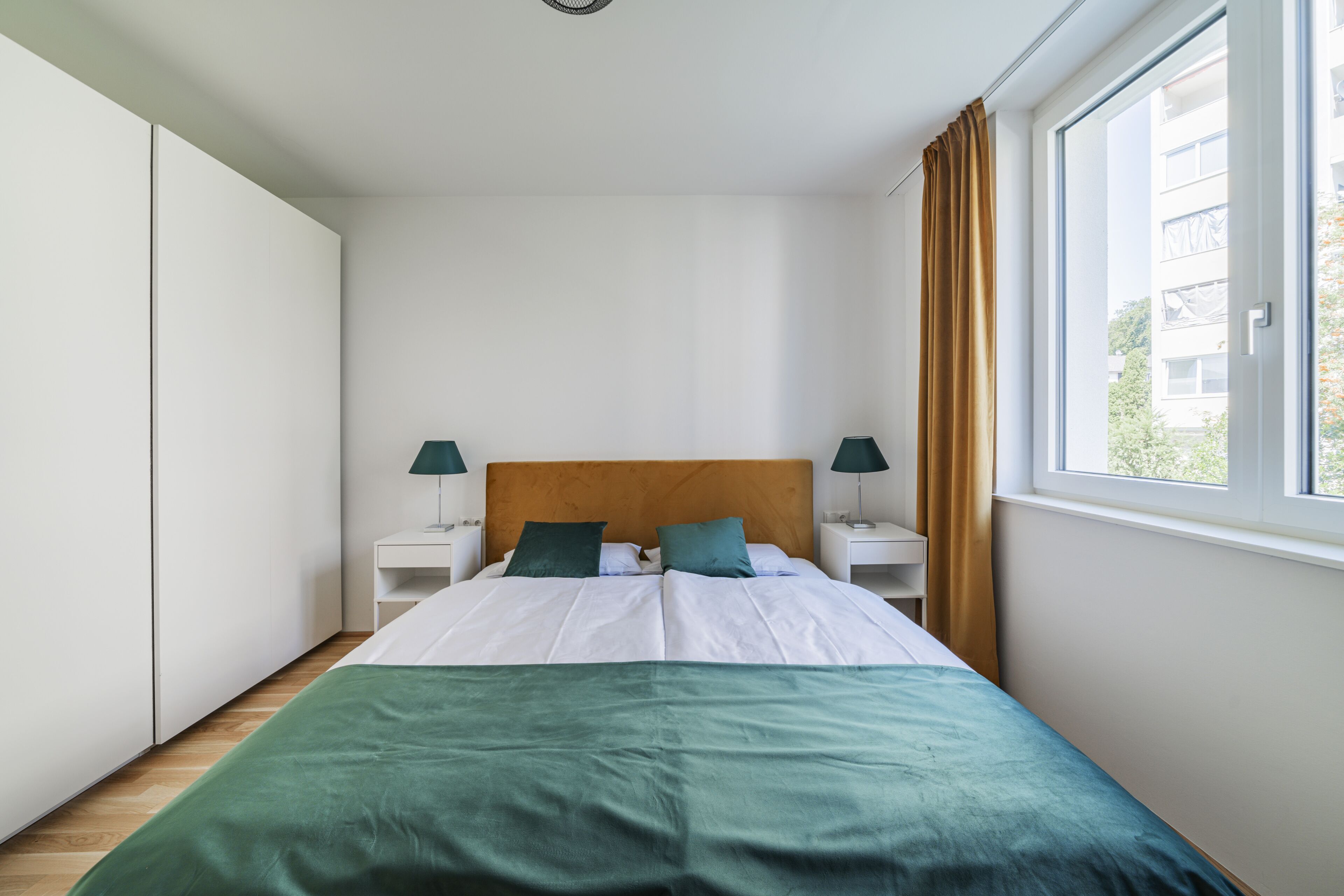 Un dormitorio con una cama, dos mesitas de noche con lámparas, una ventana con cortinas y un armario.