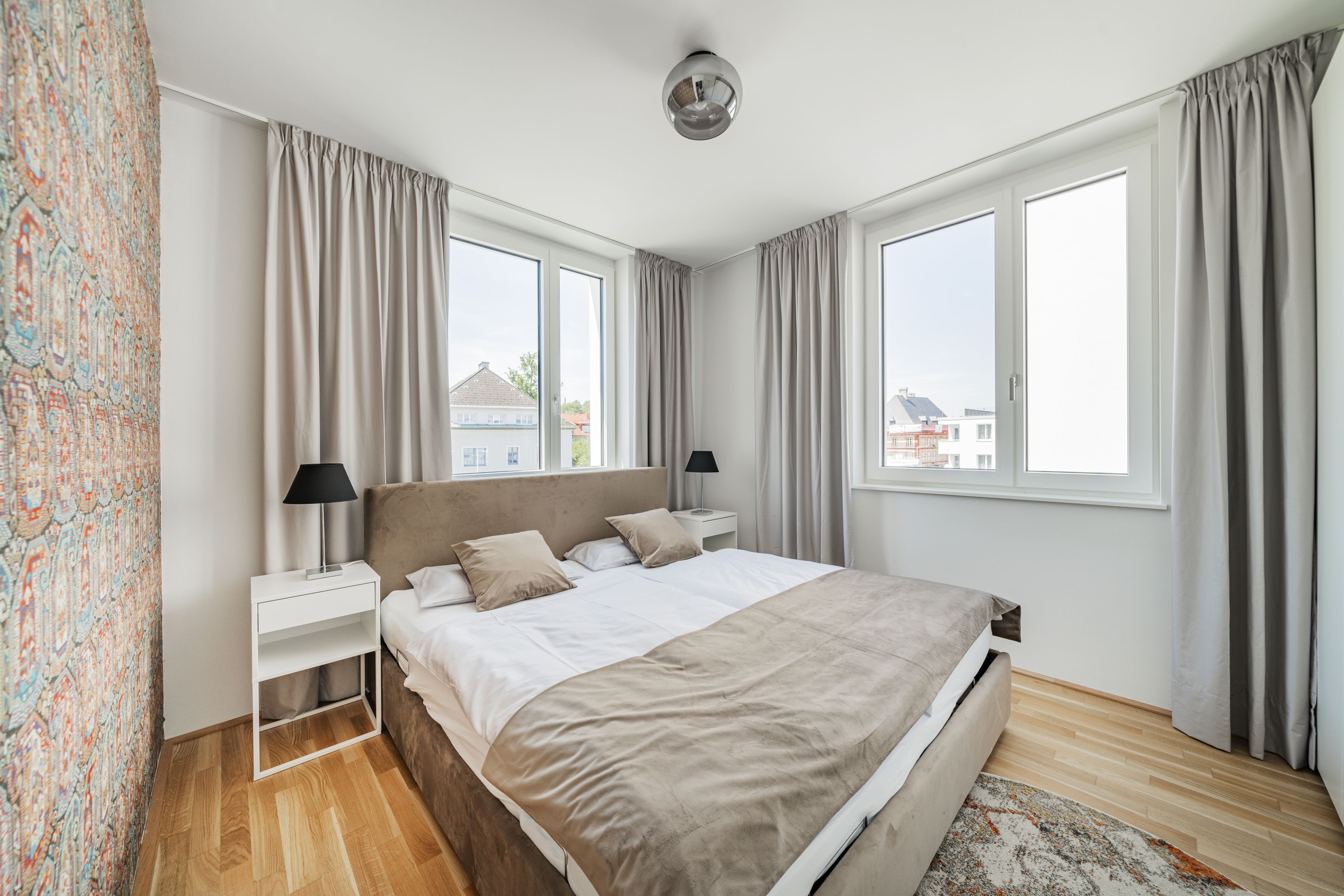 A modern bedroom with a large bed, two bedside tables, a nightstand, a ceiling light, and a view of the outside.