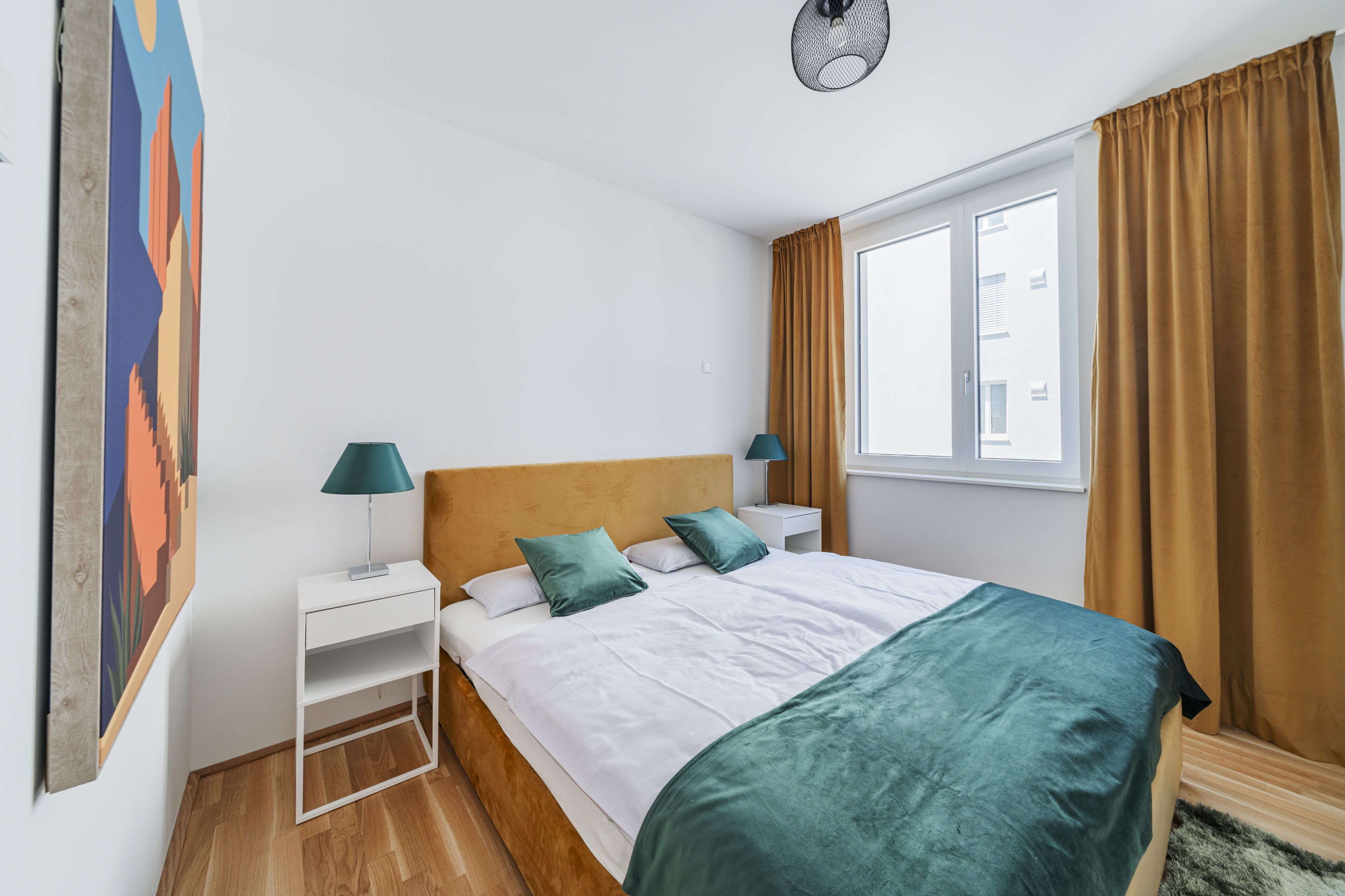 A bedroom with a bed, two bedside tables, a nightstand, a window with curtains, and a wall-mounted artwork.