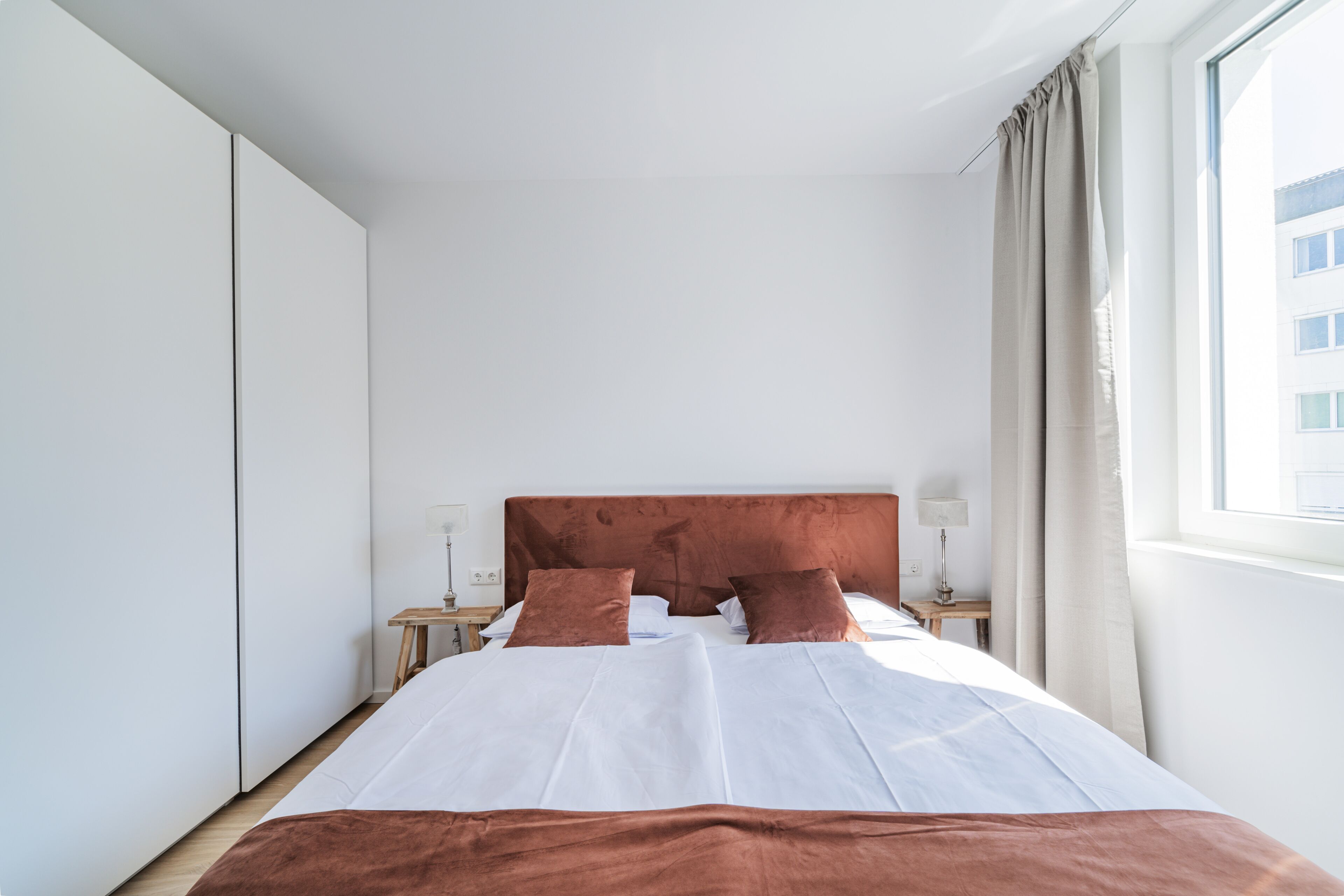 A modern hotel room with a large bed, a nightstand, a lamp, and a view of a building outside.