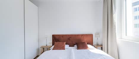 A modern hotel room with a large bed, a nightstand, a lamp, and a view of a building outside.