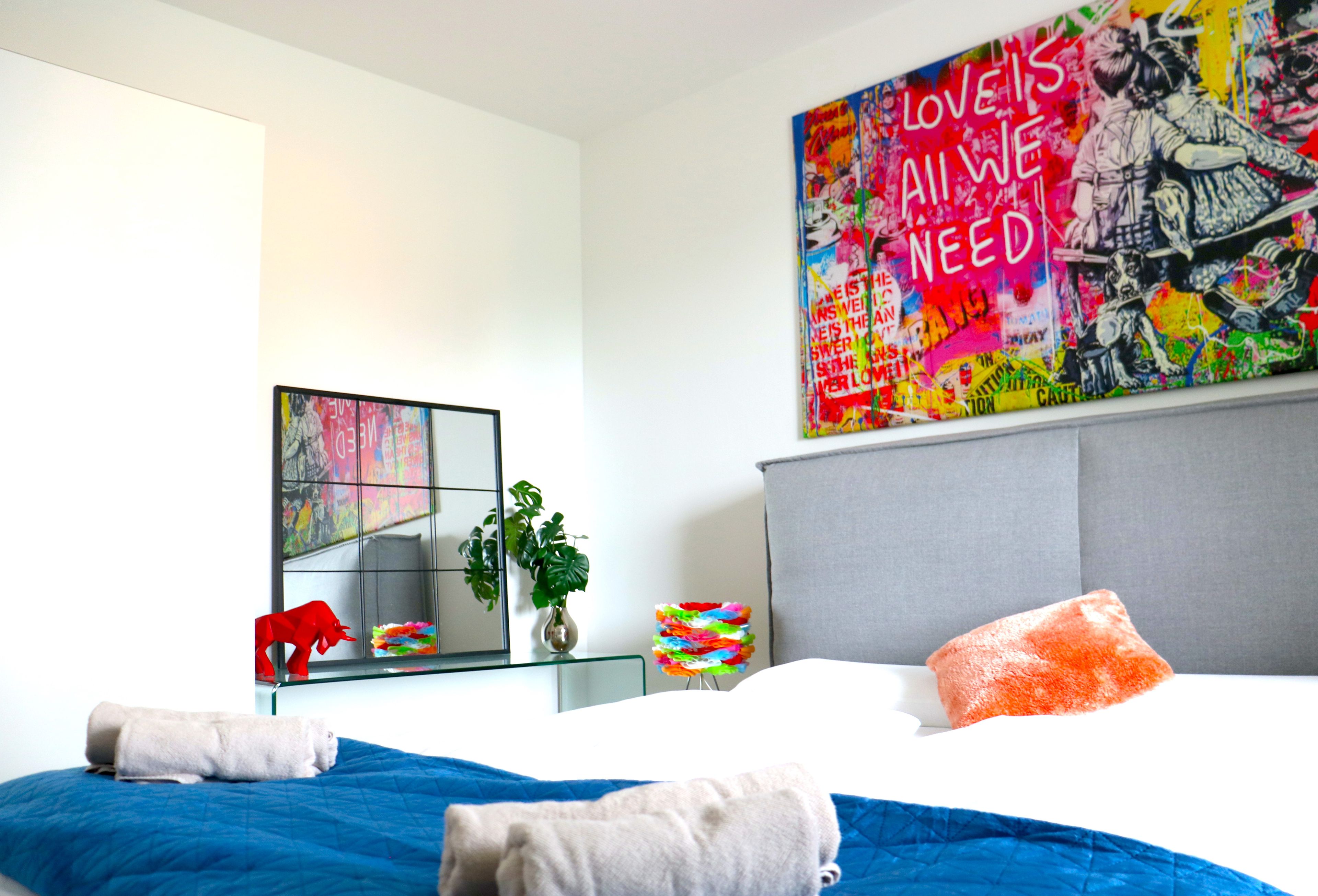 Habitación de hotel con cama, un espejo grande, una obra de arte llamativa en la pared, una planta en maceta y una mesita de noche con un florero y un objeto de colores.