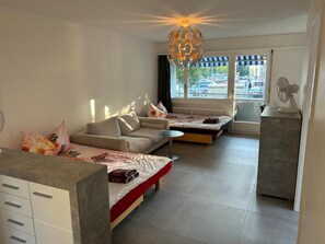 A modern living room with a sofa, a coffee table, and a dining table. There is a window with a view of the street and a decorative ceiling light. - Hanna Apartment Interlaken (Matten bei Interlaken)