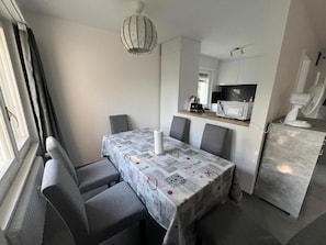 A compact dining area with a table set for four, a modern kitchen with appliances, and a large window providing natural light. - Hanna Apartment Interlaken (Matten bei Interlaken)
