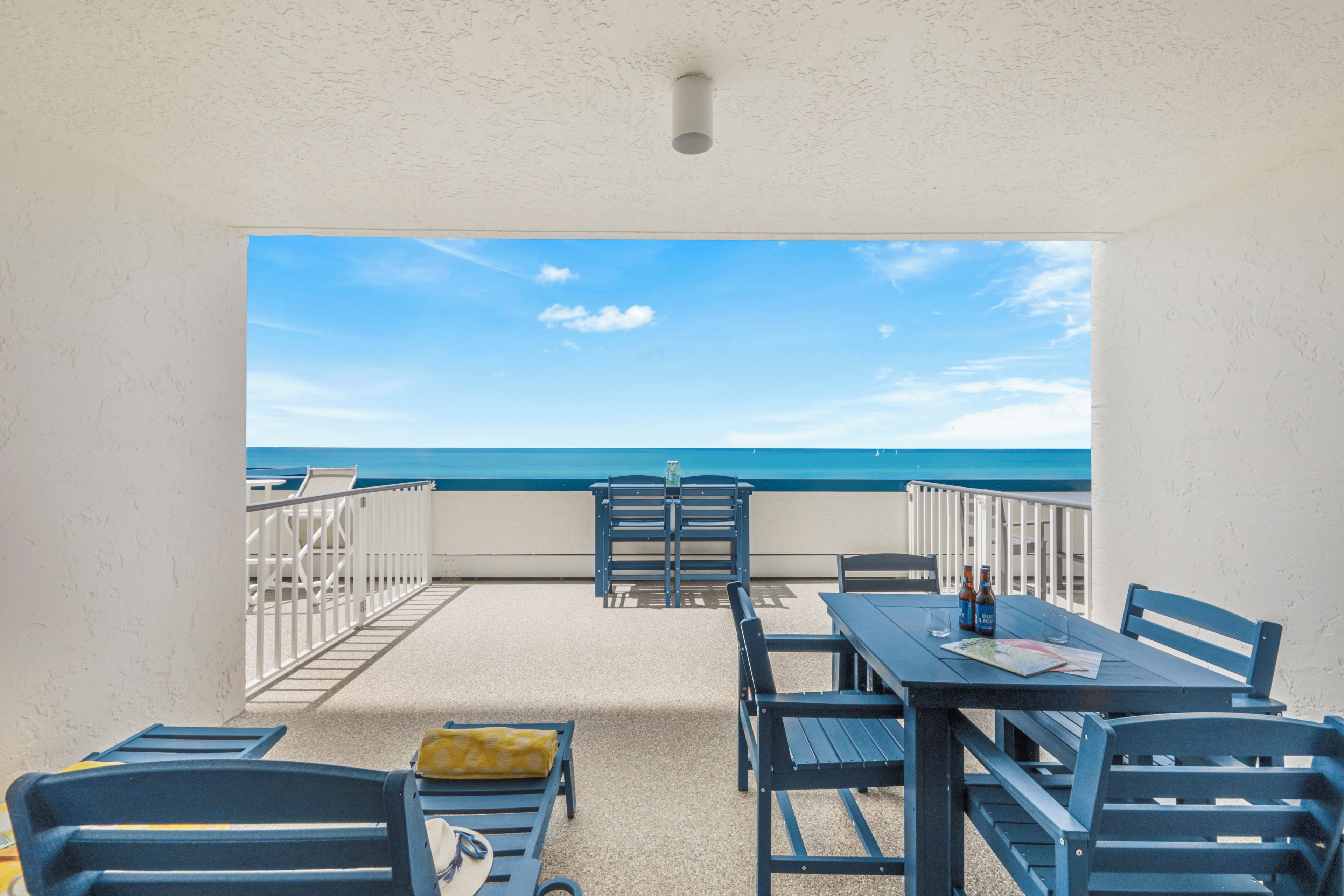 A balcony with a view of the sea, furnished with blue chairs and a table, and a white railing.