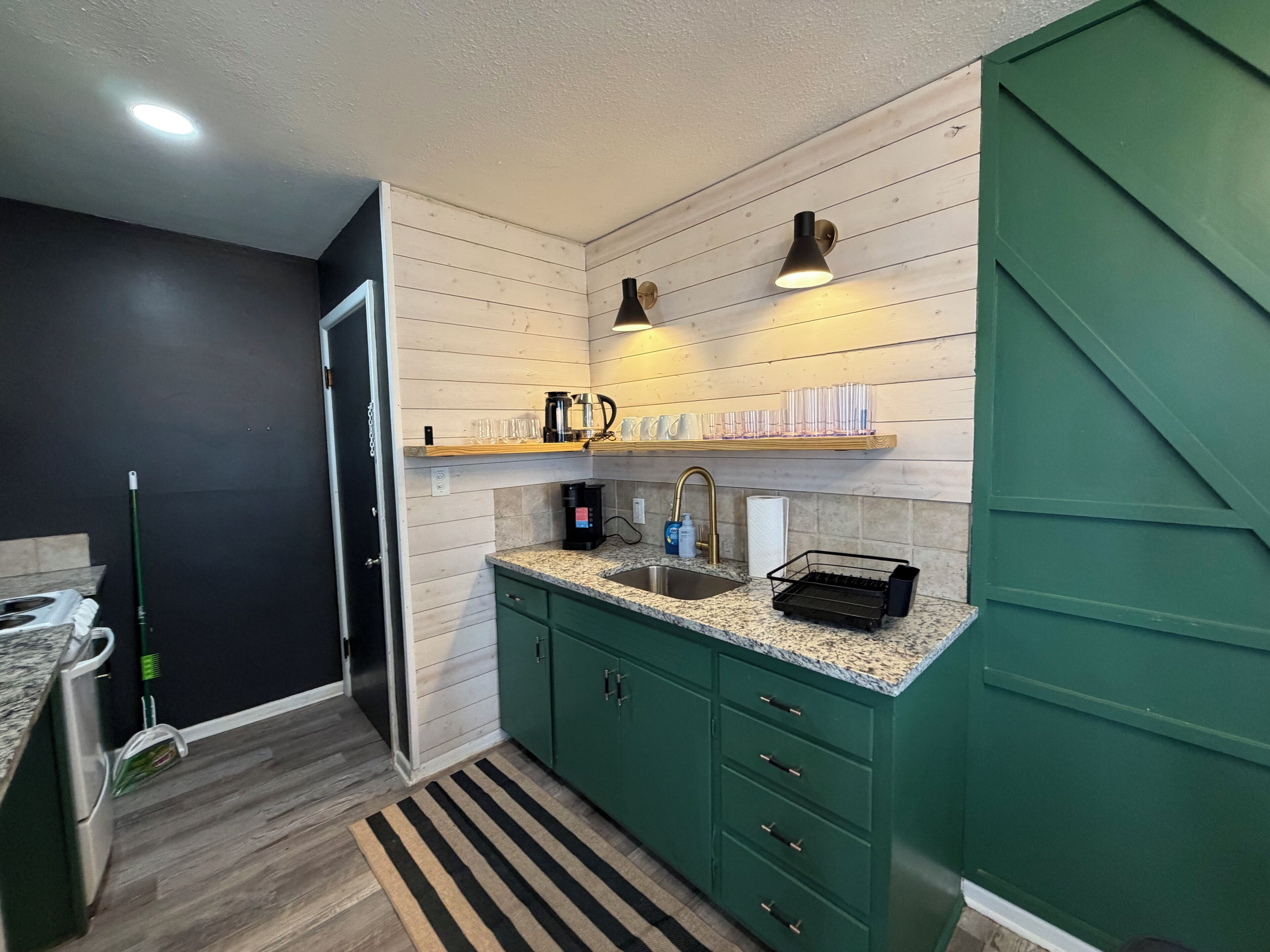 A modern kitchen with green cabinets, a granite countertop, and a built-in sink. There are shelves above the counter holding glasses and a coffee maker.