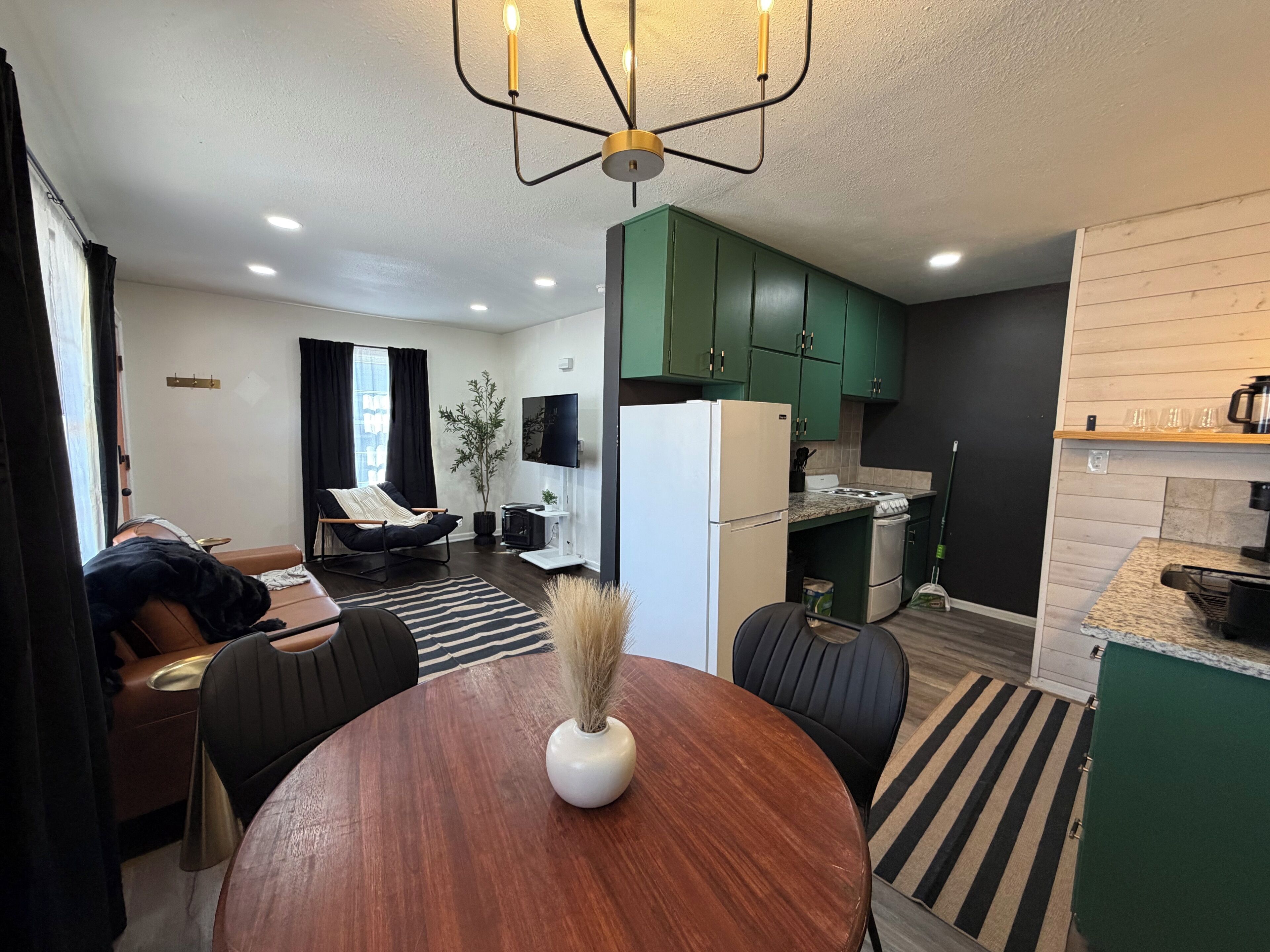 A modern living room with a dining table, a sofa, and a TV. There is a kitchen with green cabinets and a refrigerator.
