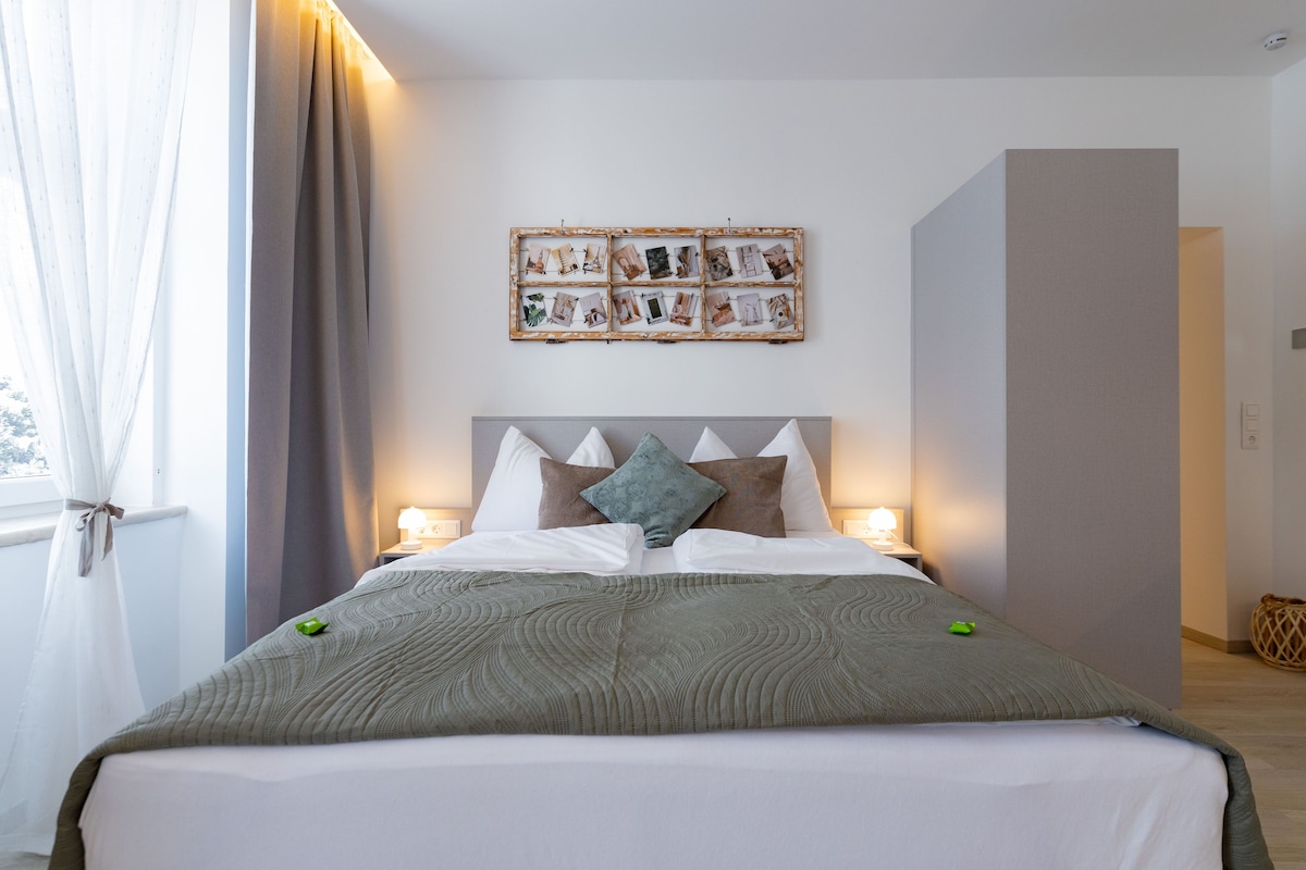 A neatly made bed with a grey quilt, a headboard with multiple pillows, bedside lamps, and a wall-mounted photo collage.