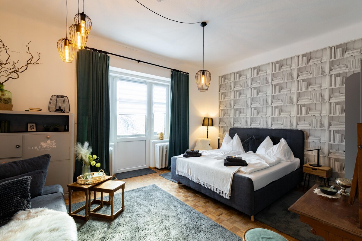 A modern bedroom with a large bed, a bedside table, a lamp, a bookshelf, and a window with curtains.