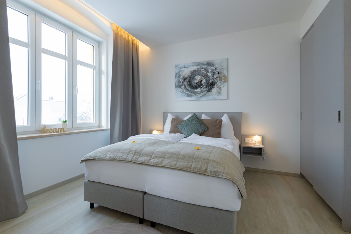 A bedroom with a bed, bedside lamps, a window with curtains, and a wall-mounted abstract painting.